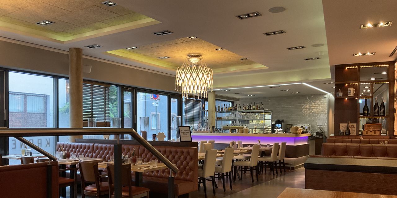 Moderner, heller Gastraum eines Restaurants mit braunen Lederbänken, Holztiischen und einer zentralen Bar, die lila beleuchtet ist. Ein auffälliger, kronleuchterartiger Lichtschirm hängt in der Mitte des Raumes.